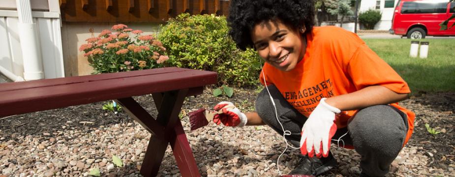 A female student in an orange t-shirt is squatting down as she paints a picnic bench a dark red color.  A can of paint in the foreground and a paint brush is in her hand. 