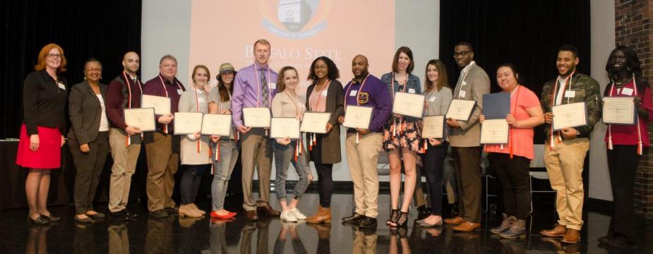 16 people are standing on a stage in front of a projection of the buffalo state logo. 14 of the 16 are holding certificates
