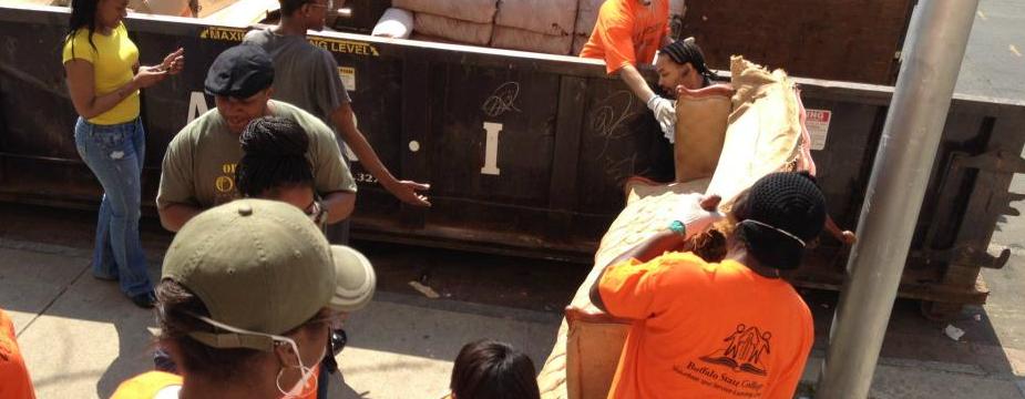 There are 10 Volunteers. 3 volunteers are loading furniture into a large dumpster on the side walk.