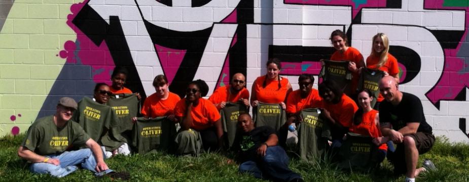 14 men and women wearing green or orange colored shirts are sitting in front of a colorful piece of artwork on the side of a building which reads OLIVER.
