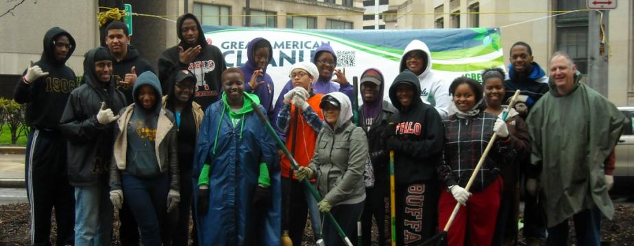 18 men and women are standing in front of a banner. A few of the people are holding rakes and brooms. 
